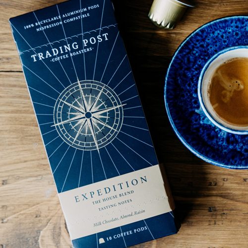 Elegant coffee packaging design for a specialty roaster, showing the Trading Post Coffee Roasters “Expedition” blend box alongside a rich espresso in a blue cup and scattered coffee pods on a wooden surface.
