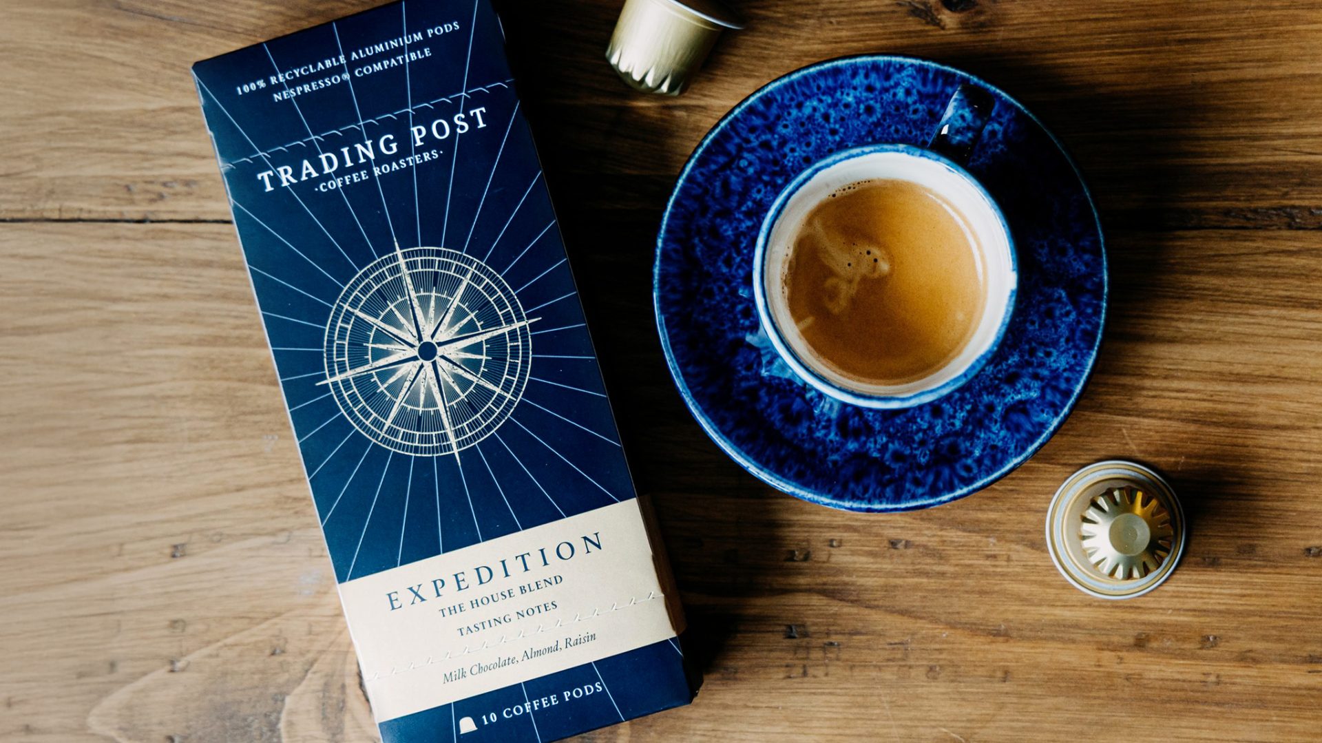 Coffee packaging design for a specialty roaster featuring a navy and cream box of Trading Post Coffee Roasters “Expedition” coffee pods beside a blue espresso cup and saucer on a wooden table.